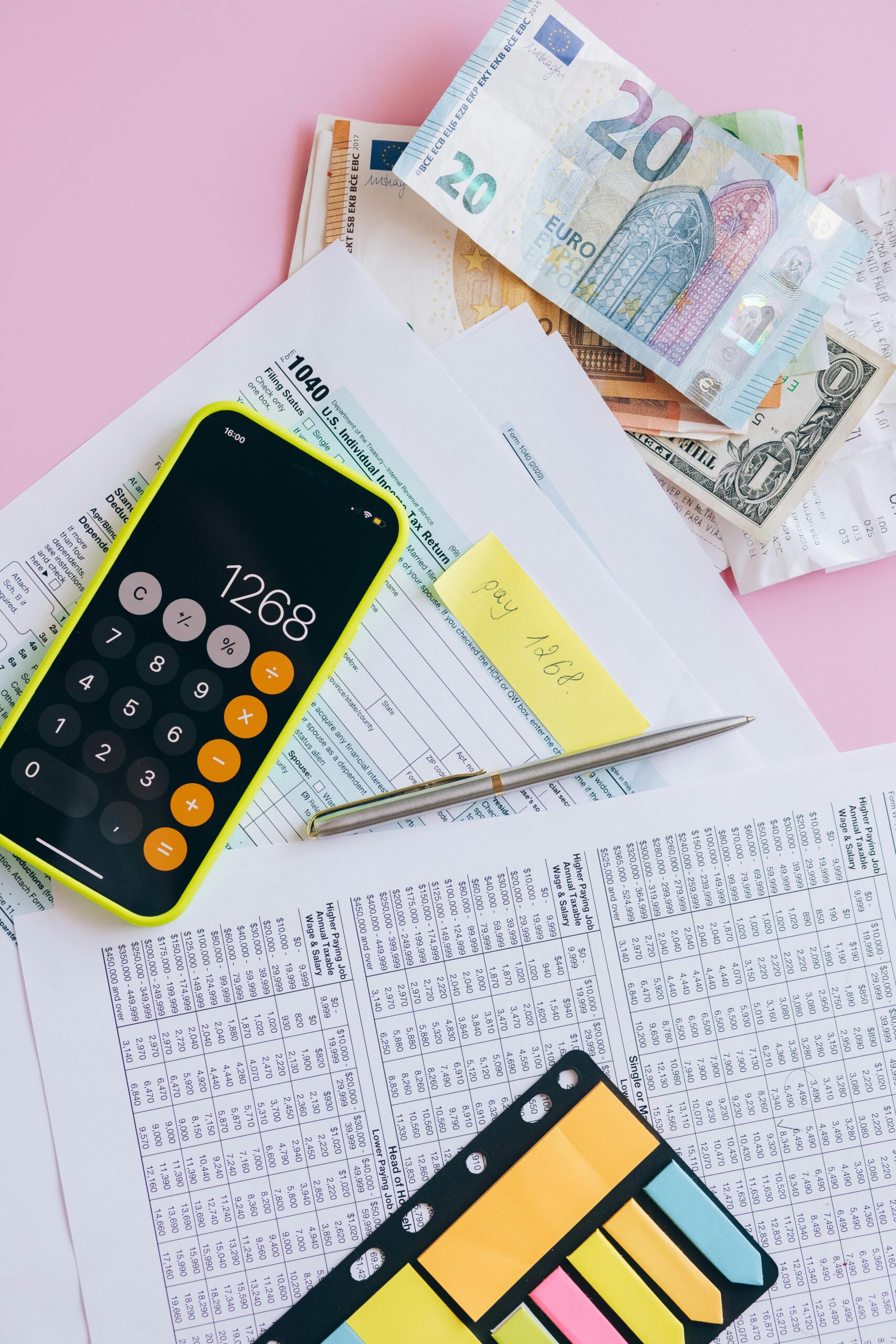 Tax documents and currency with a smartphone calculator on a pink background.